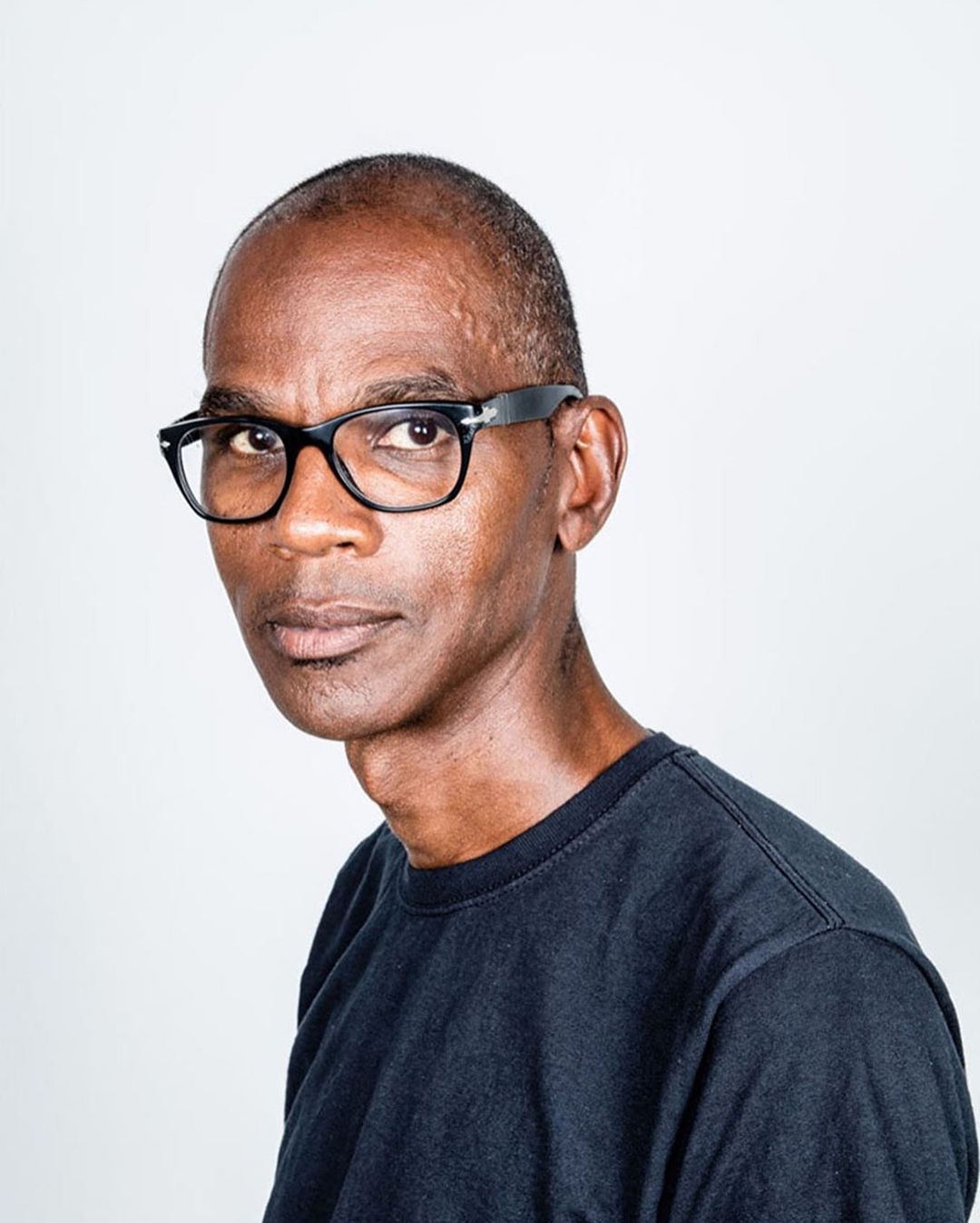 Portrait of an African American man with glasses against a light background, wearing a dark shirt, looking to the side.
