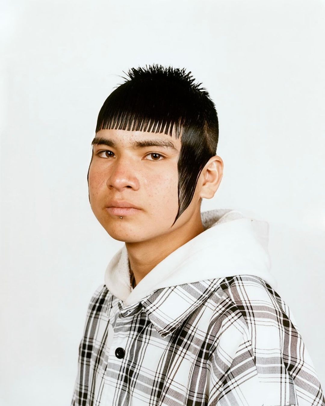 Portrait of a young man with unique black hair and a white and black plaid shirt posing indoors against a plain white background.
