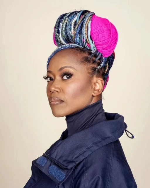 Portrait of a woman with locs styled with blue fabric and pink yarn. She wears a dark blue jacket and turtleneck in a studio setting.
