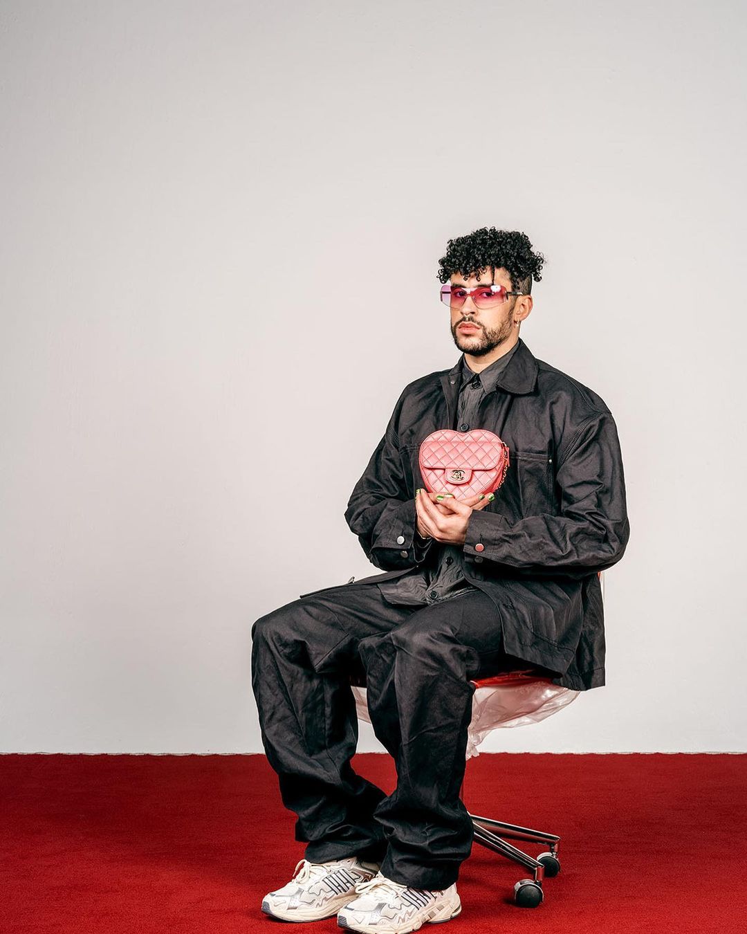 Man with pink sunglasses sits, holding a pink heart-shaped purse in a photo studio setting with a red carpet and white backdrop.