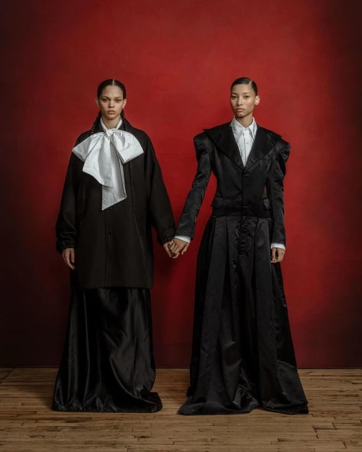 Two women in formal attire stand hand-in-hand against a red backdrop, wearing tailored black jackets, white shirts, and long black skirts.