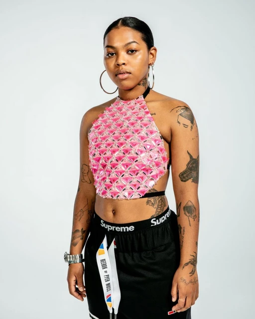 Portrait of a young adult woman with hoop earrings, black shorts, and a pink beaded crop top in a studio setting with white background.