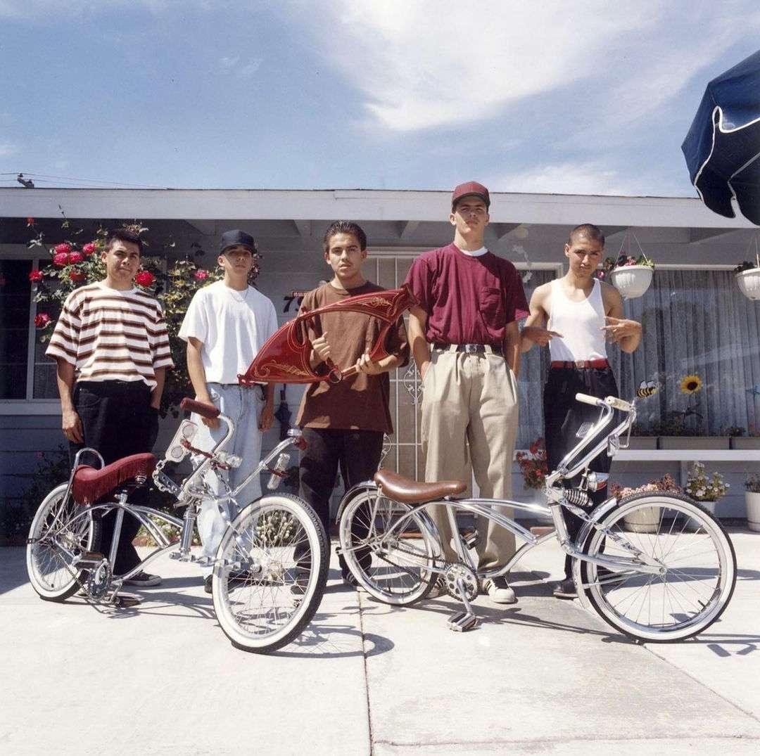 Five young men pose with customized lowrider bicycles on a sunny day in front of a light-colored house with a well-manicured lawn and rose bushes.