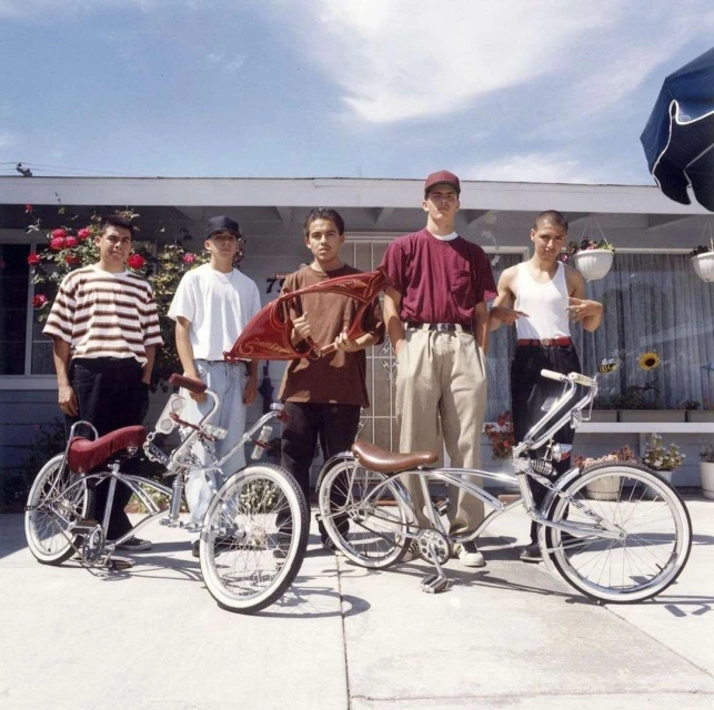 Five young men pose with customized lowrider bicycles on a sunny day in front of a light-colored house with a well-manicured lawn and rose bushes.