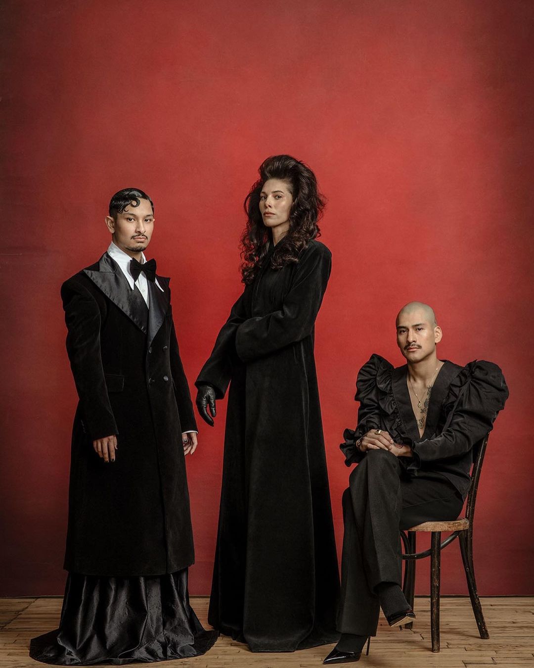 Three people in black formal wear pose against a red backdrop. One woman stands between two seated people in an indoor setting.