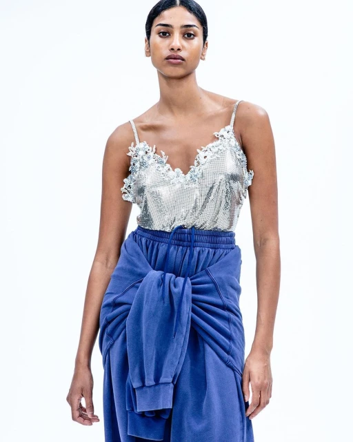 Woman in a silver sequined camisole and blue sweatpants, standing against a white background, fashion, beauty, portrait-style shot.