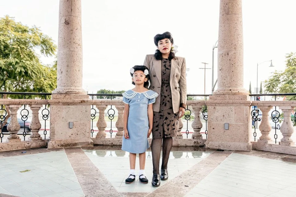 Woman and child pose in vintage outfits against a light stone rotunda with round columns and decorative iron railings on a bright day.