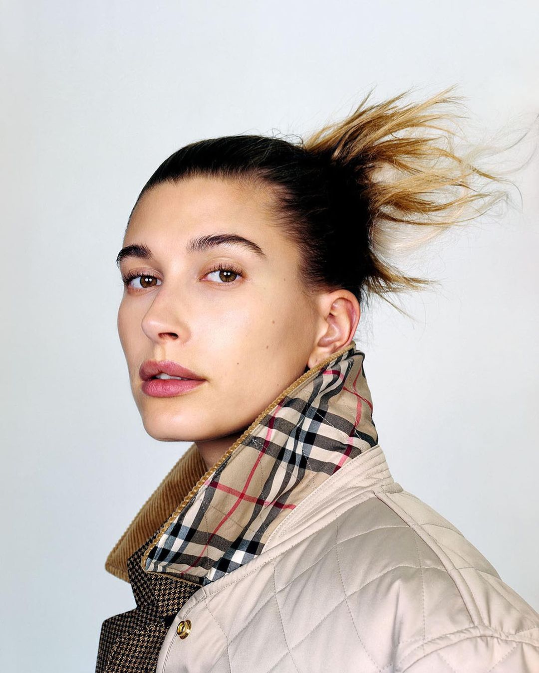 Close-up studio portrait of an adult female with brown hair, wearing a beige quilted jacket with a plaid collar against a white backdrop.