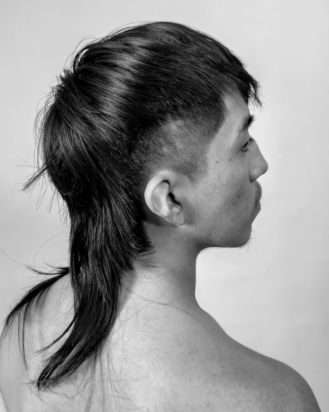 Profile portrait of an adult male with a mullet hairstyle, set against a plain background in monochrome, shoulder view.