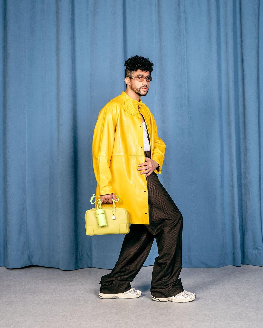 Man in a yellow jacket and dark pants, walking against a blue curtain backdrop, holding a yellow handbag.