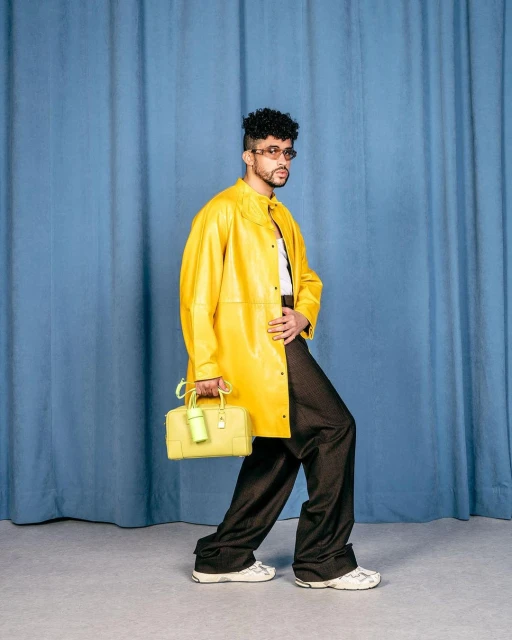 Man in a yellow jacket and dark pants, walking against a blue curtain backdrop, holding a yellow handbag.