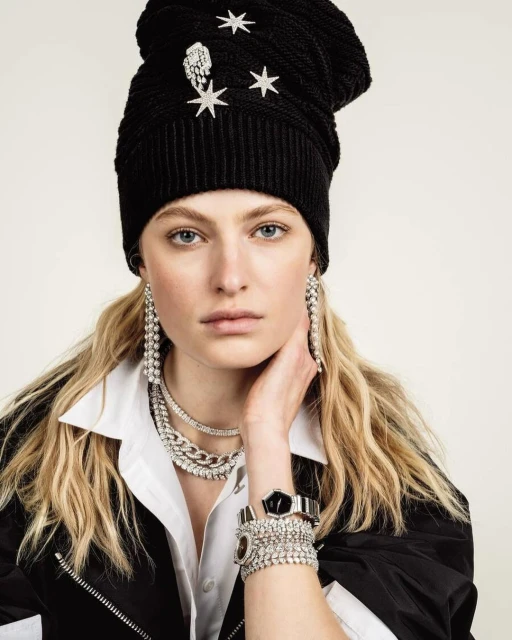 Close-up of a woman wearing a black knit hat with star embellishments, chunky diamond jewelry, a white collared shirt, and a black jacket.