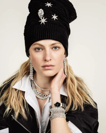 Close-up of a woman wearing a black knit hat with star embellishments, chunky diamond jewelry, a white collared shirt, and a black jacket.