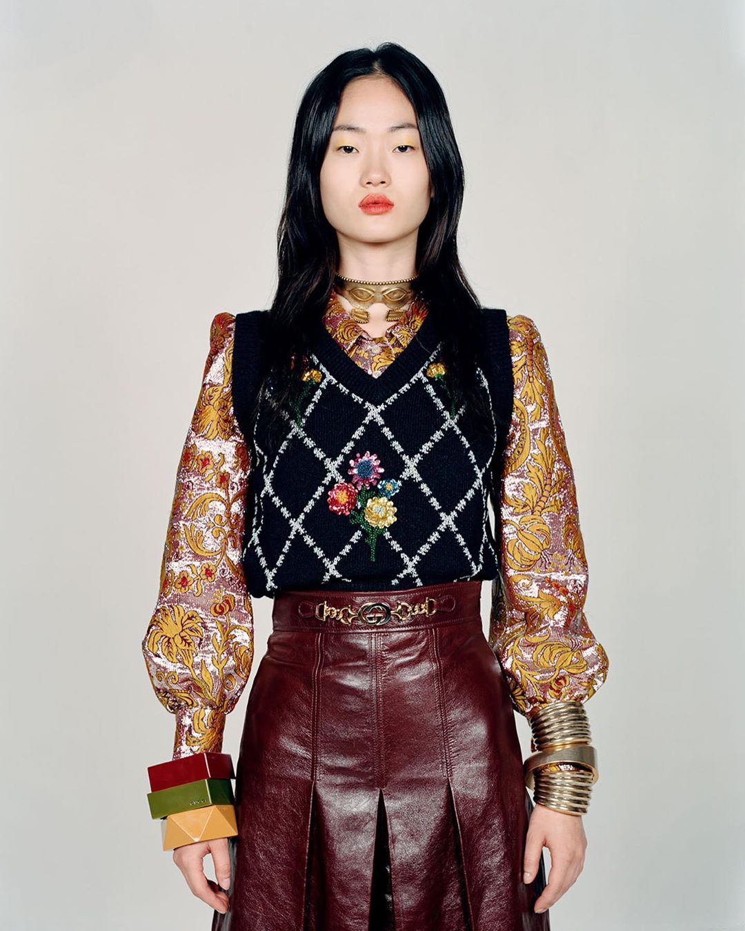 Portrait of a fashionable Asian woman wearing a floral vest, leather skirt, gold jewelry, and a patterned blouse in a studio setting.