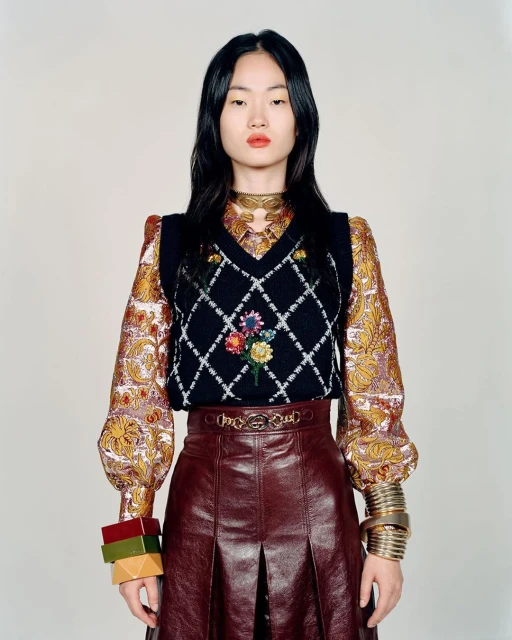 Portrait of a fashionable Asian woman wearing a floral vest, leather skirt, gold jewelry, and a patterned blouse in a studio setting.