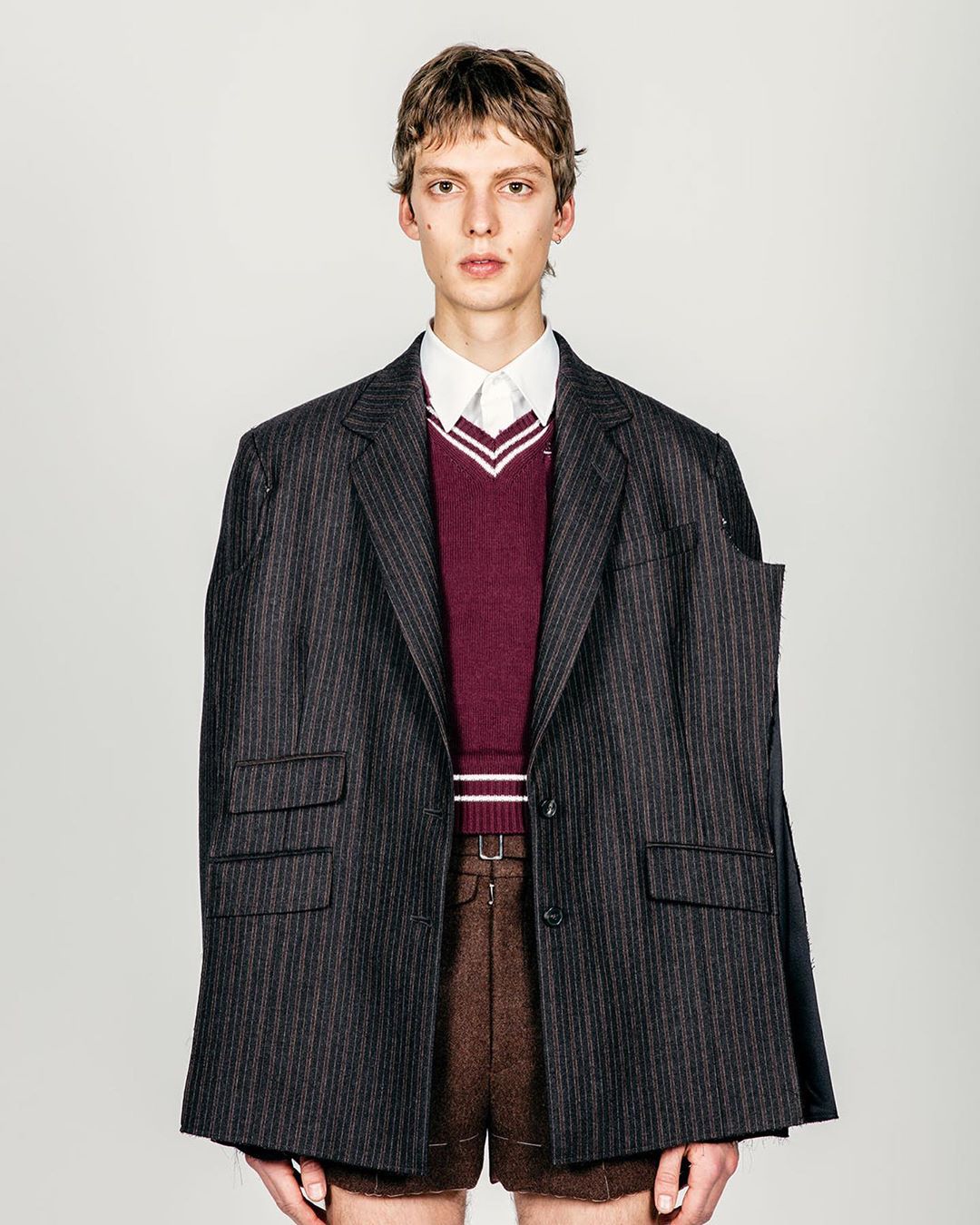 A slender adult male is photographed in a studio wearing brown shorts, a striped blazer, and a burgundy sweater with a white collared shirt.