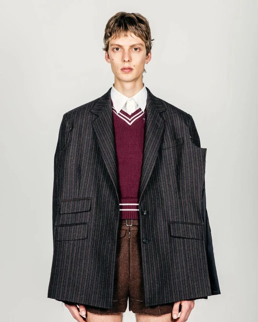 A slender adult male is photographed in a studio wearing brown shorts, a striped blazer, and a burgundy sweater with a white collared shirt.