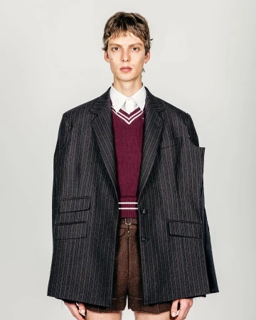 A slender adult male is photographed in a studio wearing brown shorts, a striped blazer, and a burgundy sweater with a white collared shirt.