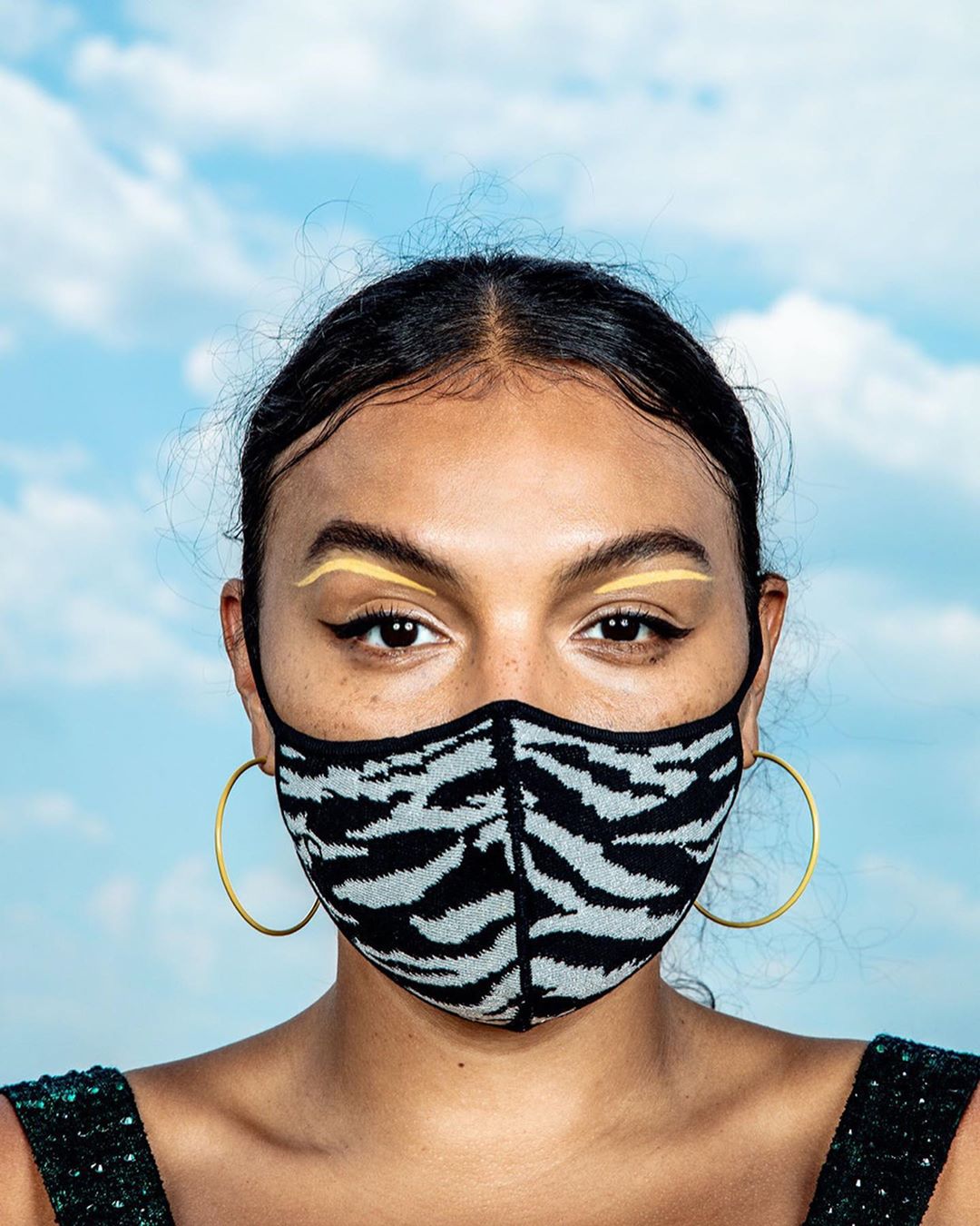 Close-up of a woman with yellow eyeliner and a zebra-striped face mask, set against a bright sky with scattered clouds.