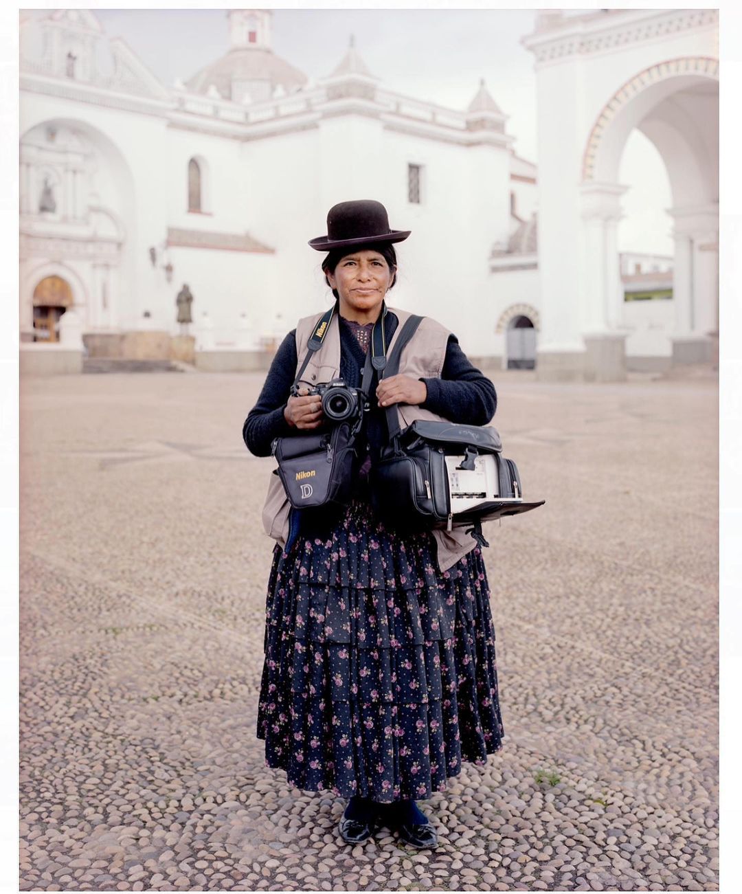 Bolivian woman photographer stands in plaza with camera gear, wearing traditional dress and bowler hat in daytime, old building in background.