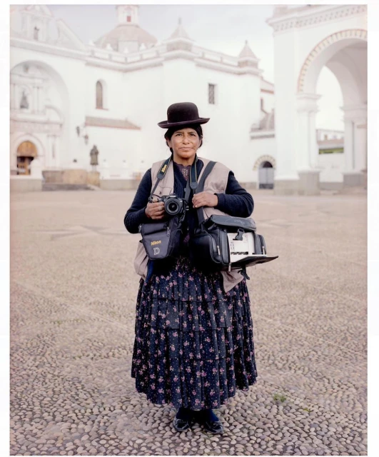 Bolivian woman photographer stands in plaza with camera gear, wearing traditional dress and bowler hat in daytime, old building in background.