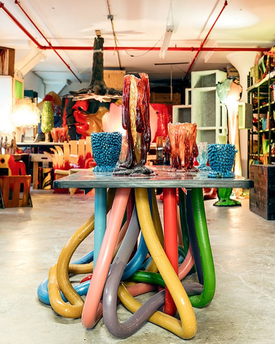Table with colorful, looping pipe legs supports unique art glass vases and bowls in an artist's studio, creating a whimsical, creative scene.