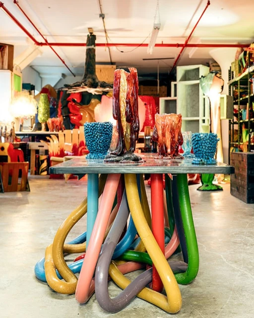 Table with colorful, looping pipe legs supports unique art glass vases and bowls in an artist's studio, creating a whimsical, creative scene.