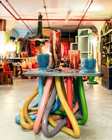 Table with colorful, looping pipe legs supports unique art glass vases and bowls in an artist's studio, creating a whimsical, creative scene.