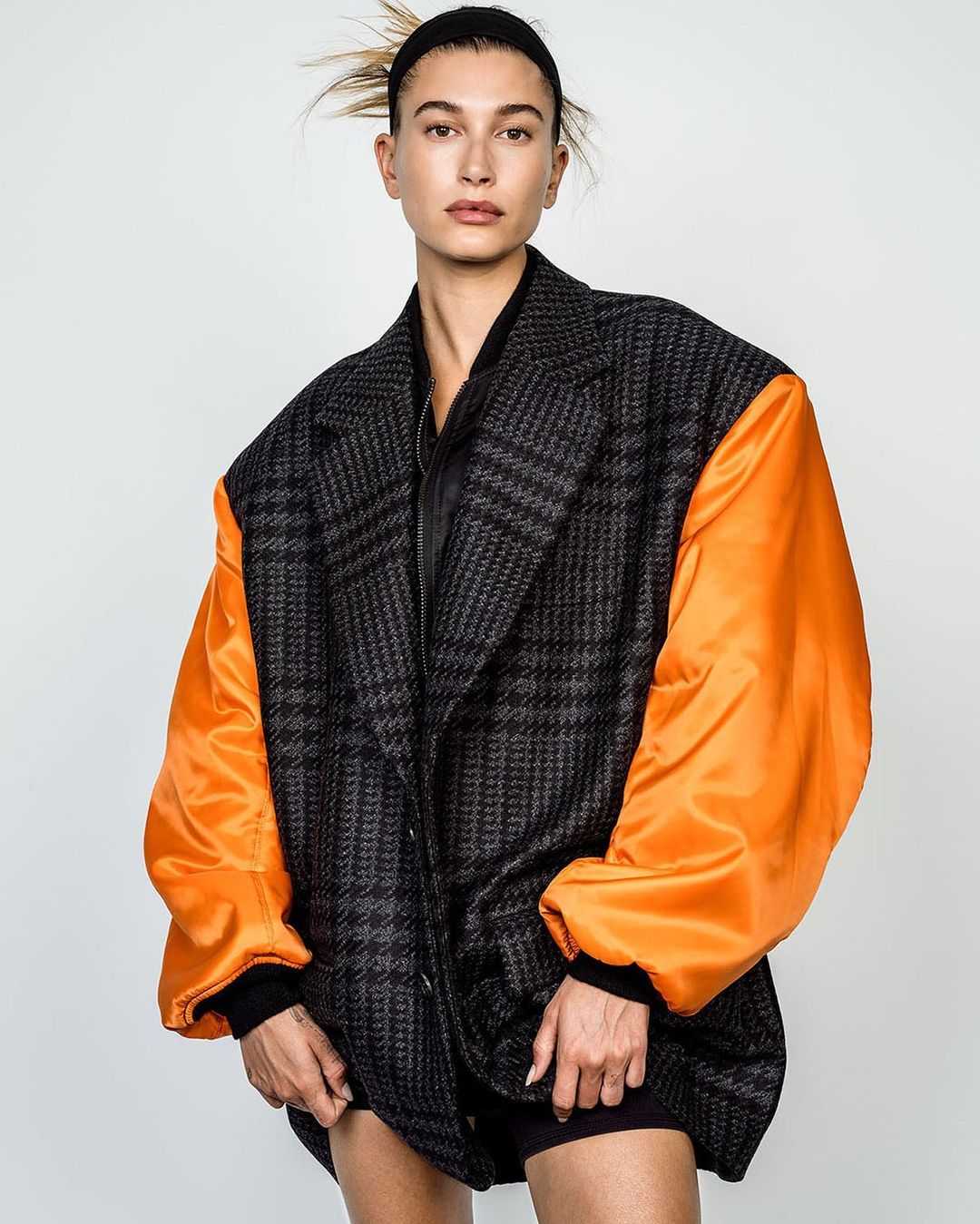 Model Hailey Bieber wears an oversized grey plaid blazer with orange sleeves in a portrait against a white backdrop in a studio.