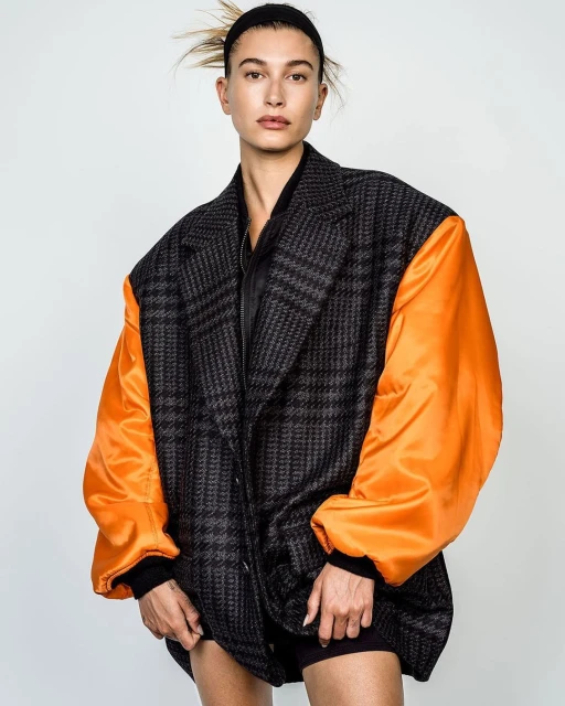 Model Hailey Bieber wears an oversized grey plaid blazer with orange sleeves in a portrait against a white backdrop in a studio.