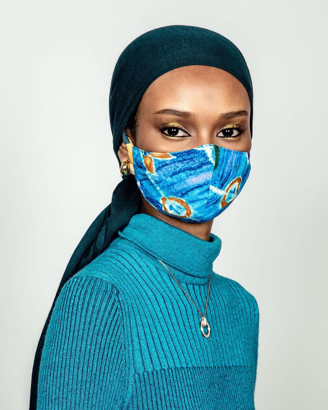 Close-up portrait of an adult female with dark hair wearing a turquoise turtleneck, headscarf, patterned face mask and jewelry on a white background.