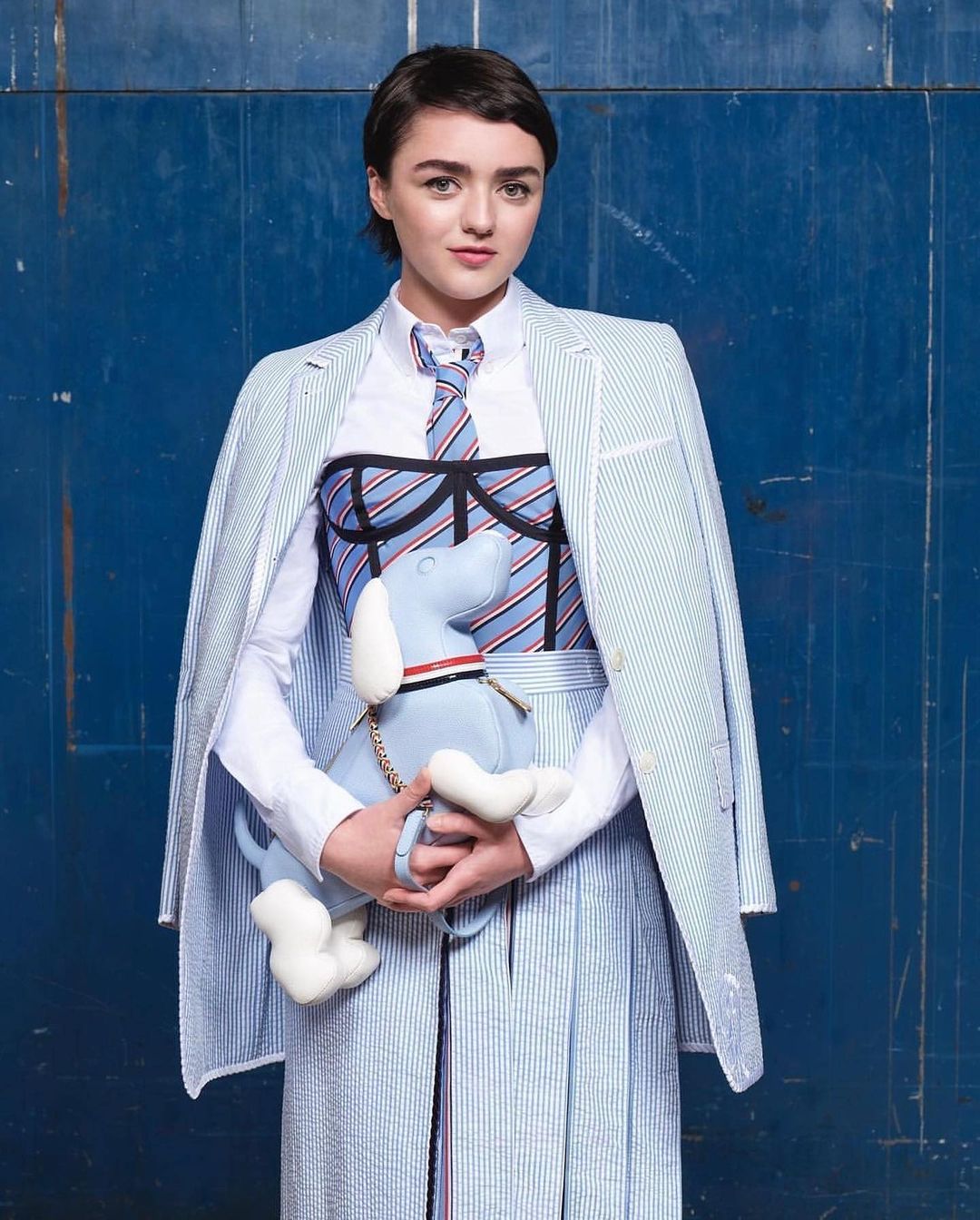 Maisie Williams in striped blazer, skirt, and tie, holding a plush dog, set against a blue backdrop in a fashion forward studio portrait.