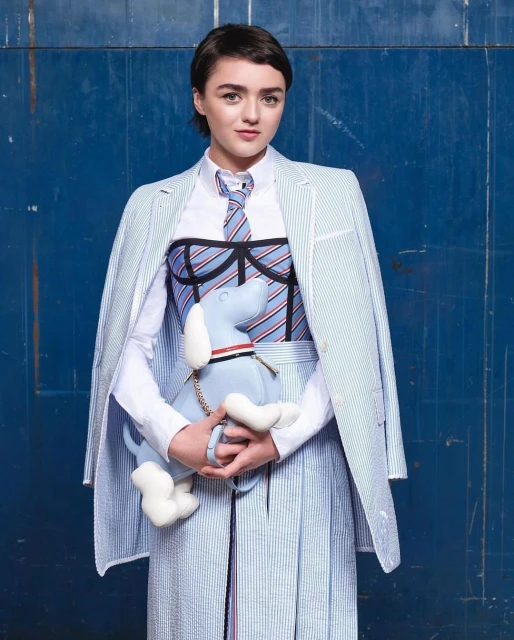 Maisie Williams in striped blazer, skirt, and tie, holding a plush dog, set against a blue backdrop in a fashion forward studio portrait.