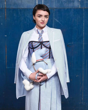 Maisie Williams in striped blazer, skirt, and tie, holding a plush dog, set against a blue backdrop in a fashion forward studio portrait.