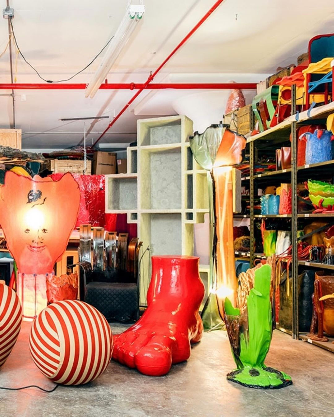 Whimsical collection of oversized red and green objects, including a giant foot and light fixtures, displayed in an eclectic warehouse setting.
