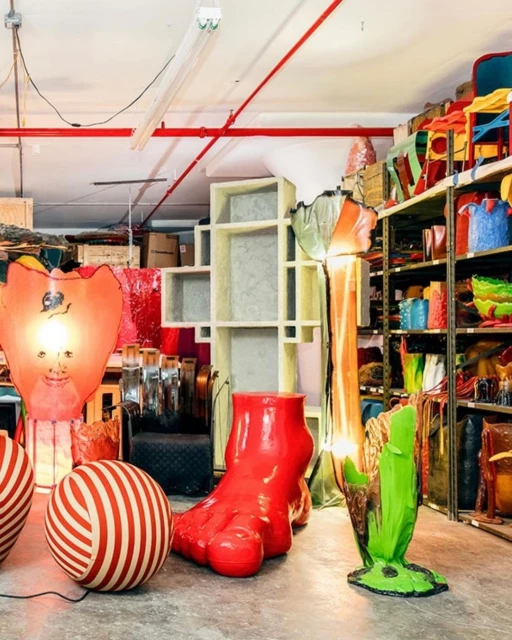Whimsical collection of oversized red and green objects, including a giant foot and light fixtures, displayed in an eclectic warehouse setting.