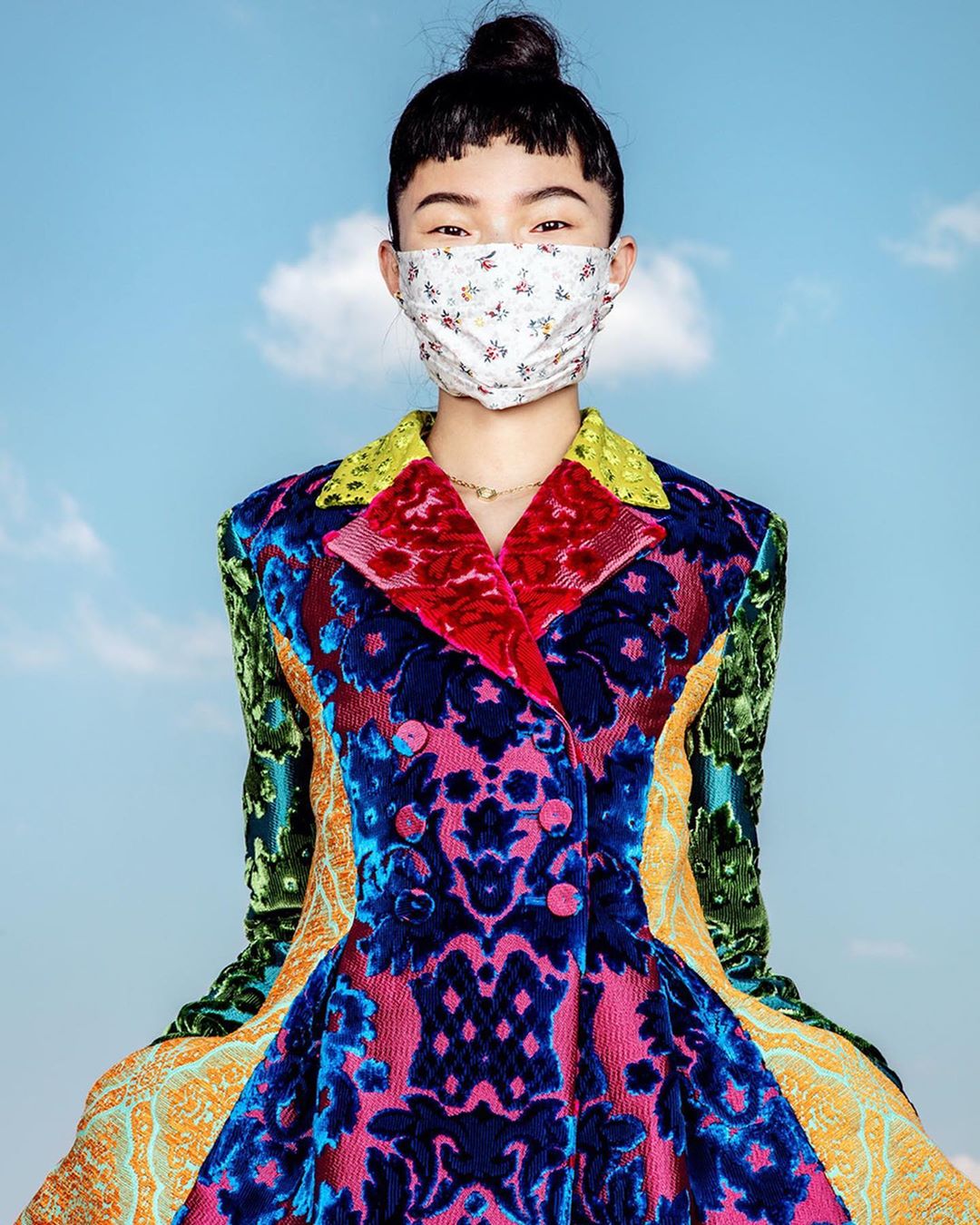 Asian woman in a colorful patterned jacket and floral face mask posing against a bright sky with soft clouds. Fashion editorial style.