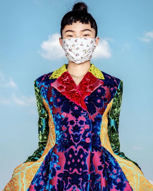 Asian woman in a colorful patterned jacket and floral face mask posing against a bright sky with soft clouds. Fashion editorial style.