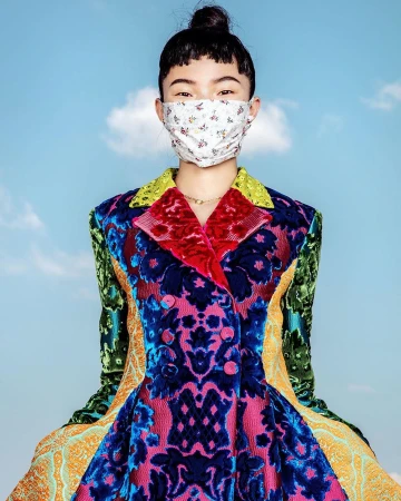 Asian woman in a colorful patterned jacket and floral face mask posing against a bright sky with soft clouds. Fashion editorial style.
