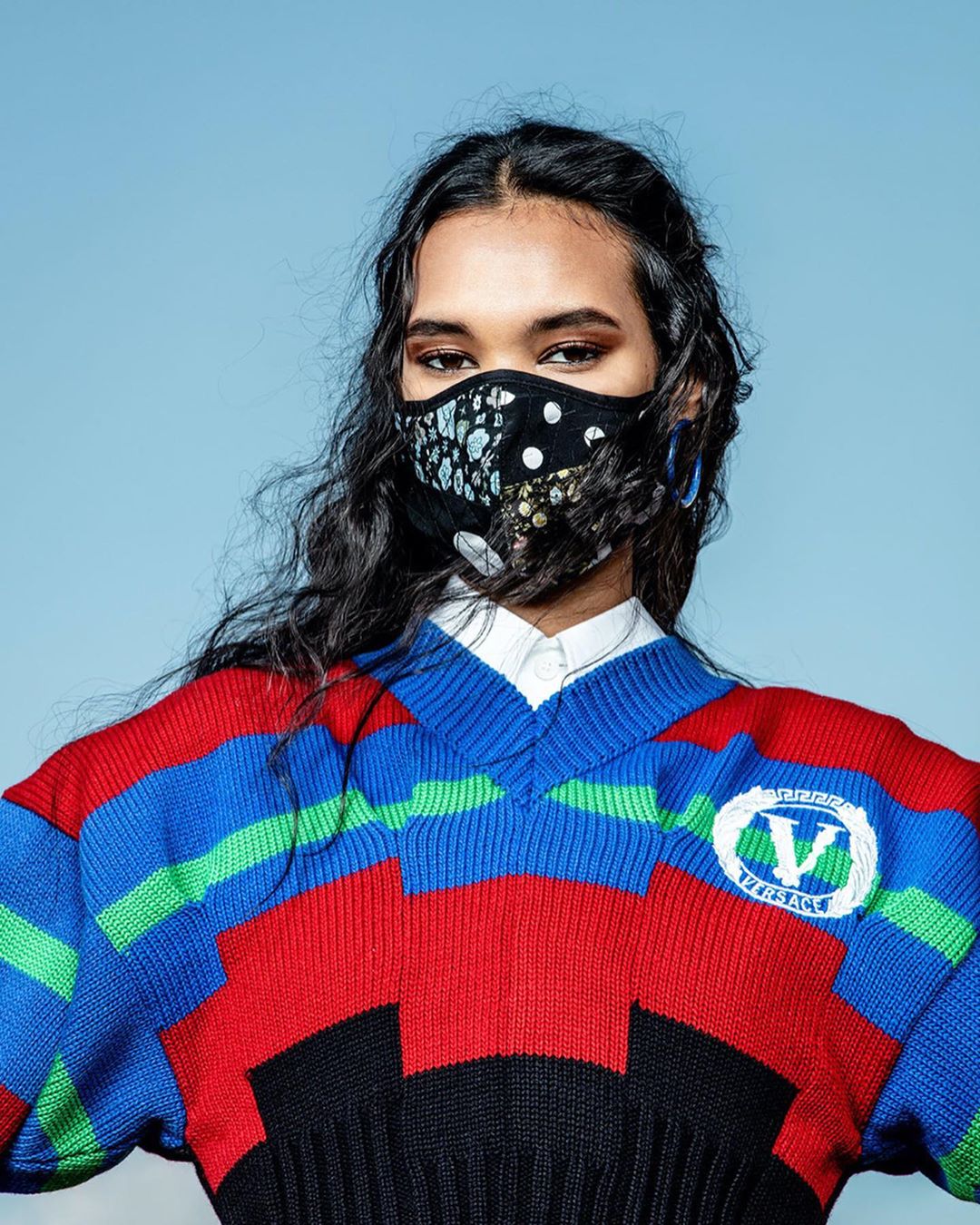A woman with dark hair wears a patterned face mask and a bright striped sweater against a blue background in a fashion-style portrait.