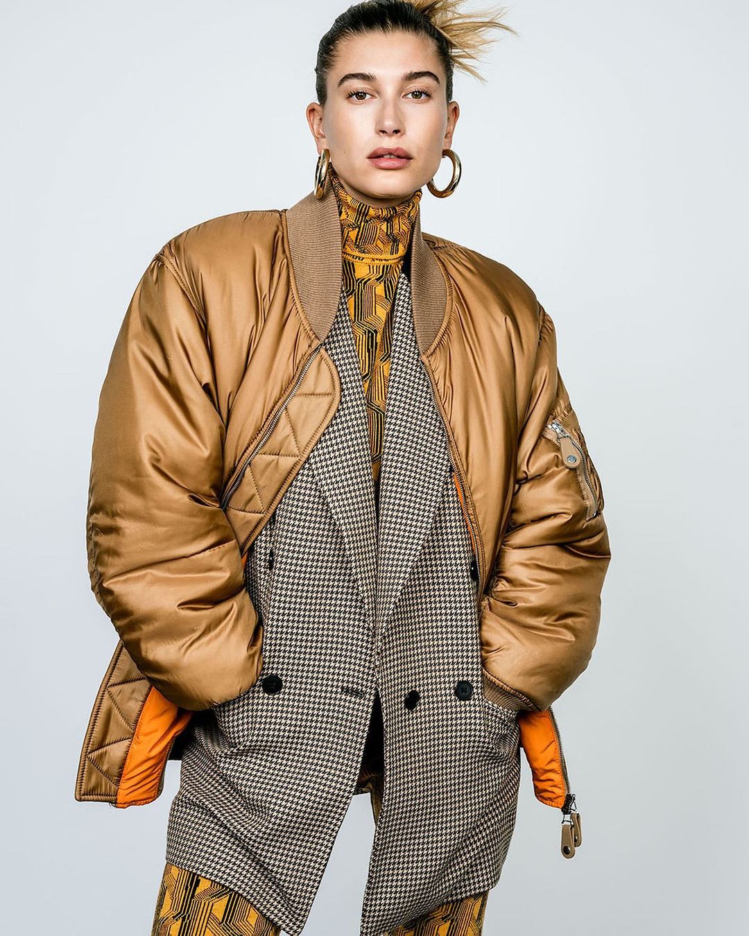 Woman in a gold bomber jacket, houndstooth blazer, and patterned turtleneck poses against a plain backdrop, showcasing a fashion-forward style.