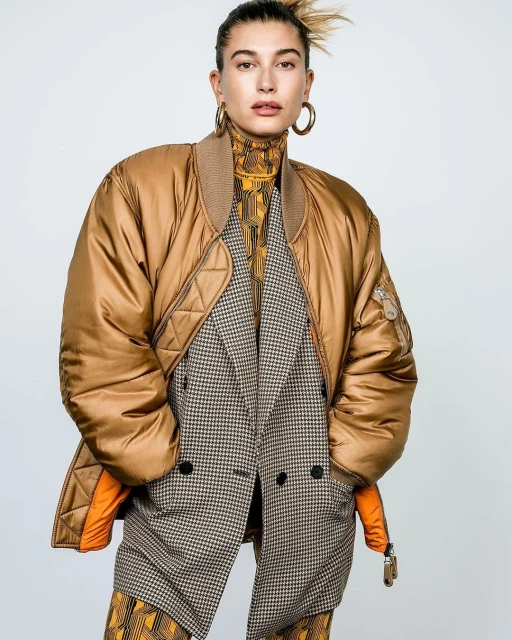 Woman in a gold bomber jacket, houndstooth blazer, and patterned turtleneck poses against a plain backdrop, showcasing a fashion-forward style.