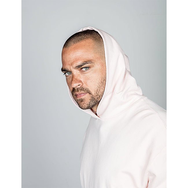 Portrait of a man with short hair and light eyes, wearing a pink hooded sweatshirt, against a light gray background in a studio.