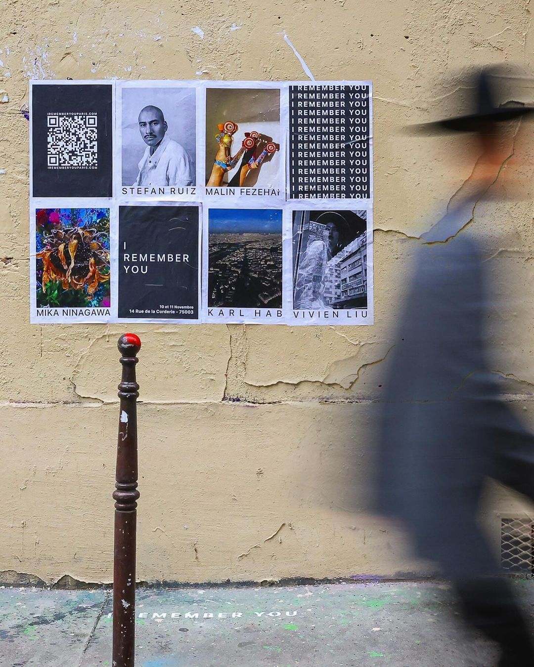 Art posters adorn an aged, textured wall, featuring names like Mika Ninagawa and Karl Hab, alongside a blurred figure walking past in motion.