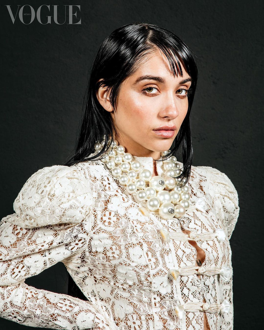 Portrait of a woman with dark hair wearing a white lace top and large pearl necklace, photographed against a dark gray background.