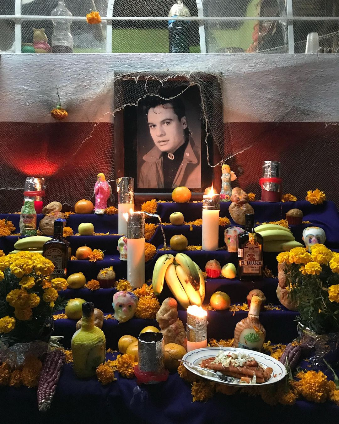 Altar for Day of the Dead with portrait, candles, marigolds, fruit, and food offerings on tiered shelves against a white wall.