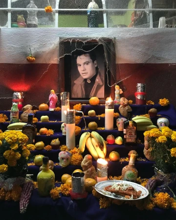 Altar for Day of the Dead with portrait, candles, marigolds, fruit, and food offerings on tiered shelves against a white wall.