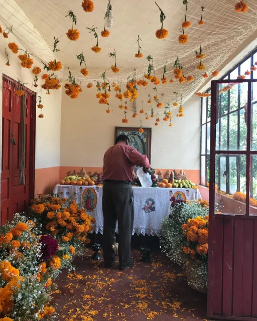 Day of the Dead altar with flowers, fruit, and a man arranging offerings in an indoor setting with bright orange marigolds.
