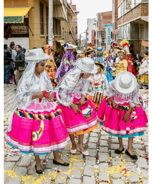 Group of young girls in matching traditional dresses and hats parade down a cobblestone street strewn with flower petals during a daytime festival.