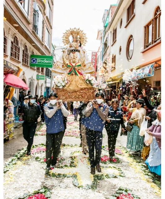 Procession through a city street with people wearing face masks, carrying an ornate statue, and walking on a flower-covered path.